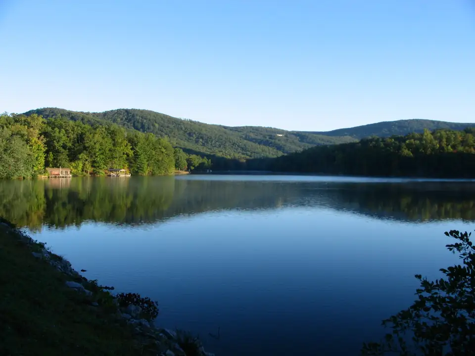 A still lake reflecting forested ridges under a pale sky, with two small boathouses on the far shore.