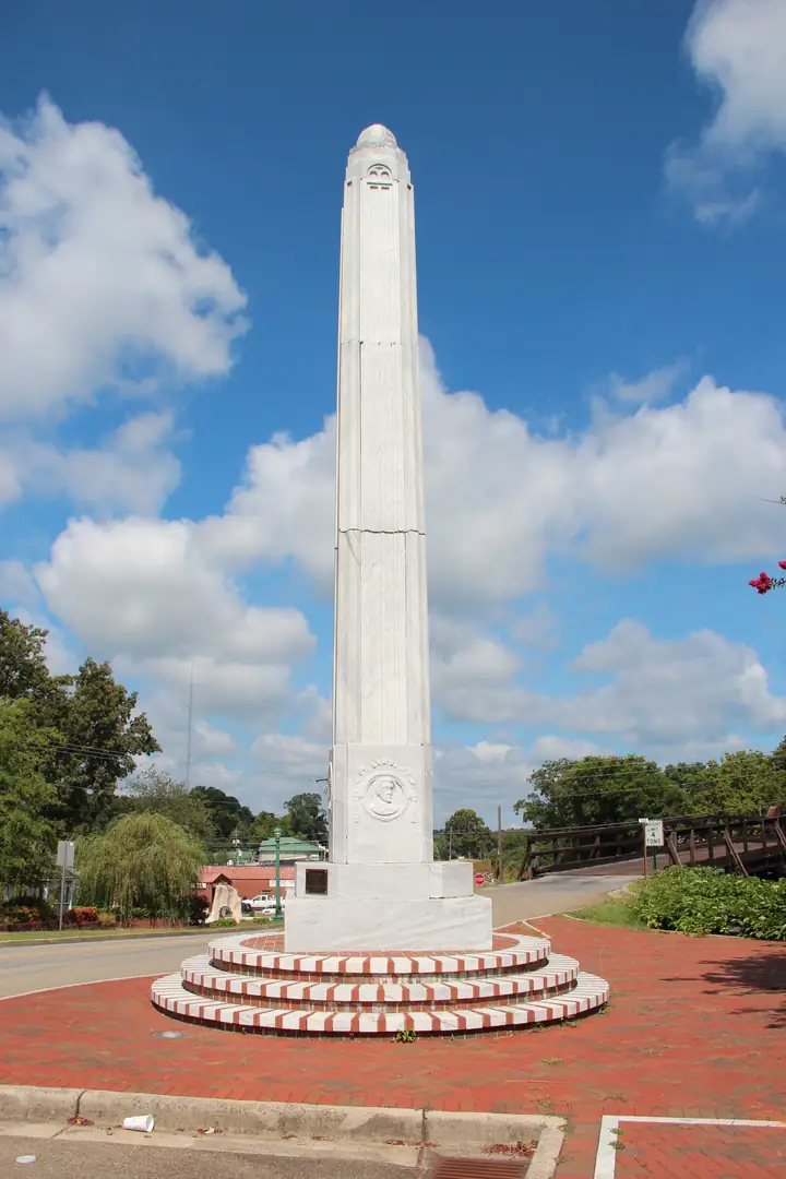 A tall white marble obelisk with a carved portrait medallion, standing on brick tiers in a paved roundabout.
