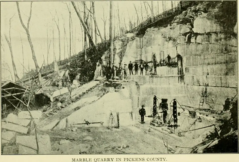 Black-and-white photo of men standing on the stepped white ledges of an open marble quarry cut into a wooded hill.