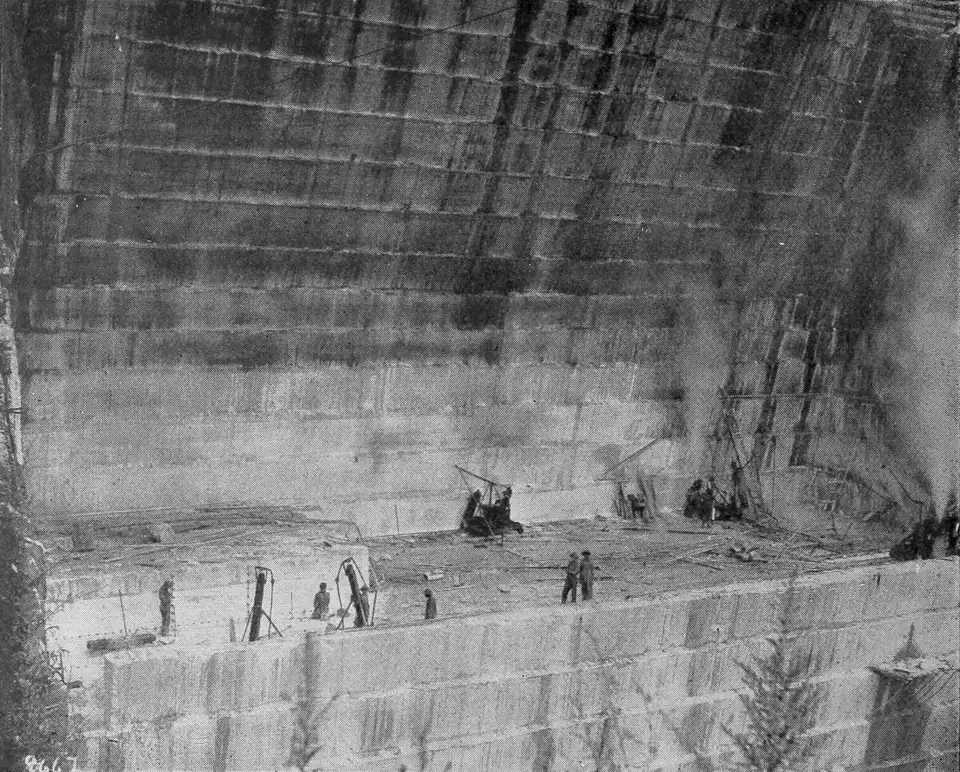 Black-and-white photo looking down into a deep marble quarry with stepped walls, workers, machines and rising steam.