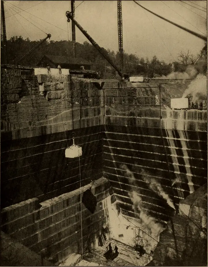 Sepia photo of a marble block hanging from a derrick cable above a very deep, square-cut quarry pit.