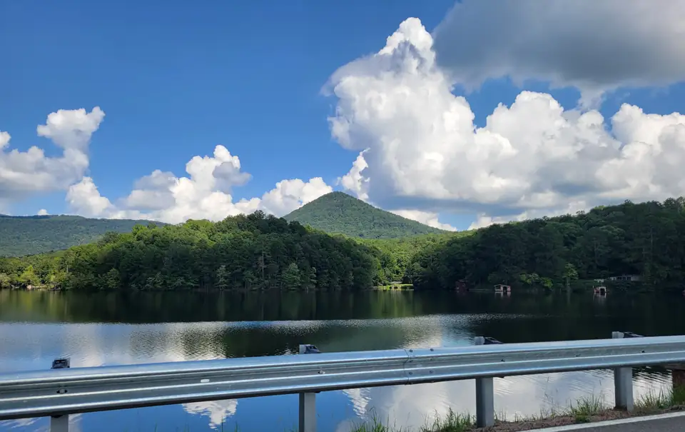 A pointed green peak behind a forested shore, with tall white summer clouds reflected in a dark lake.