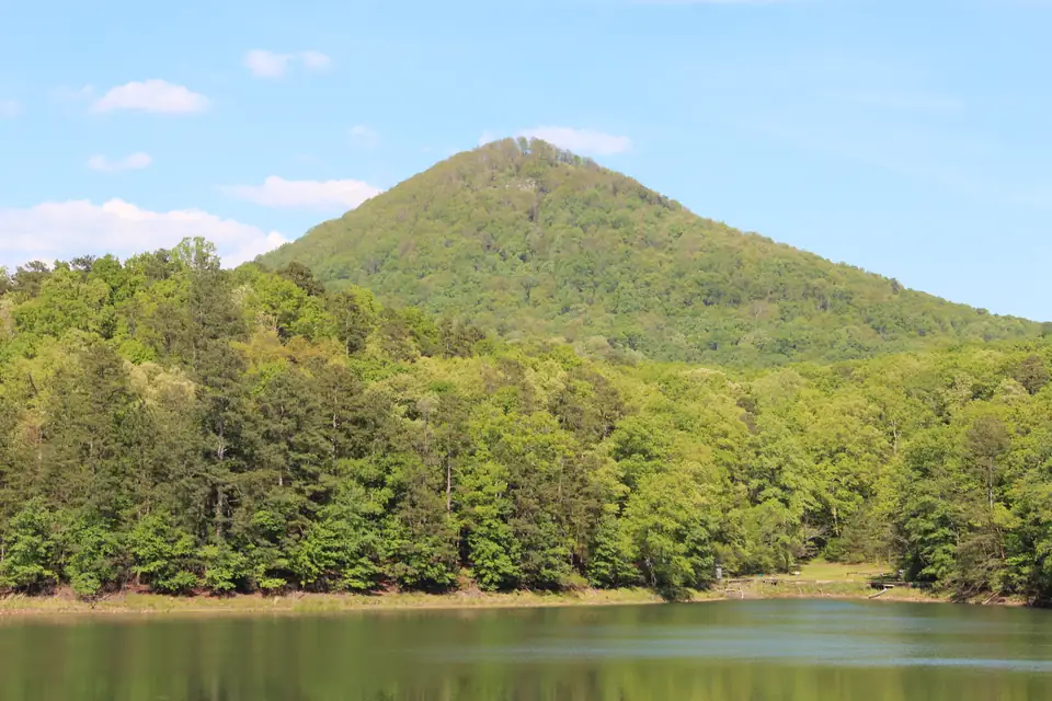 A cone-shaped wooded mountain rising behind a bright green shoreline and calm green lake water.