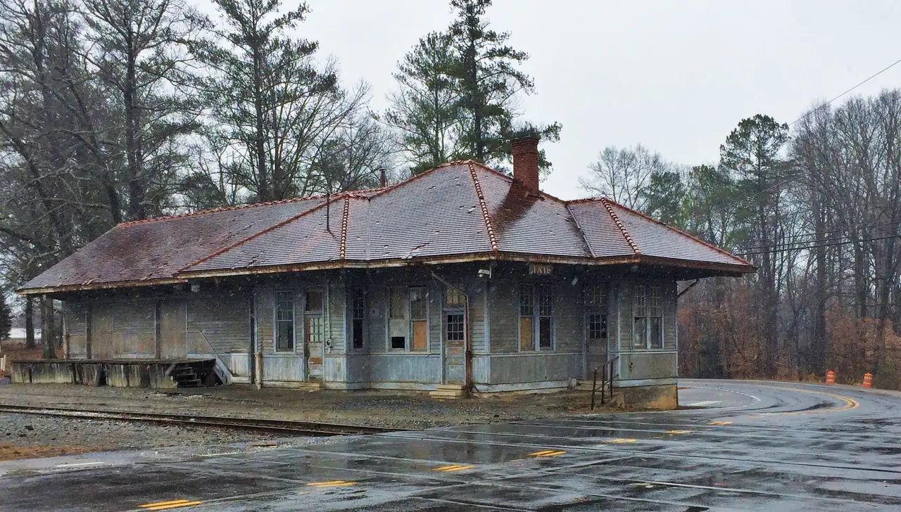 A long, low clapboard depot with a red tile roof beside a railroad crossing, under falling snow.
