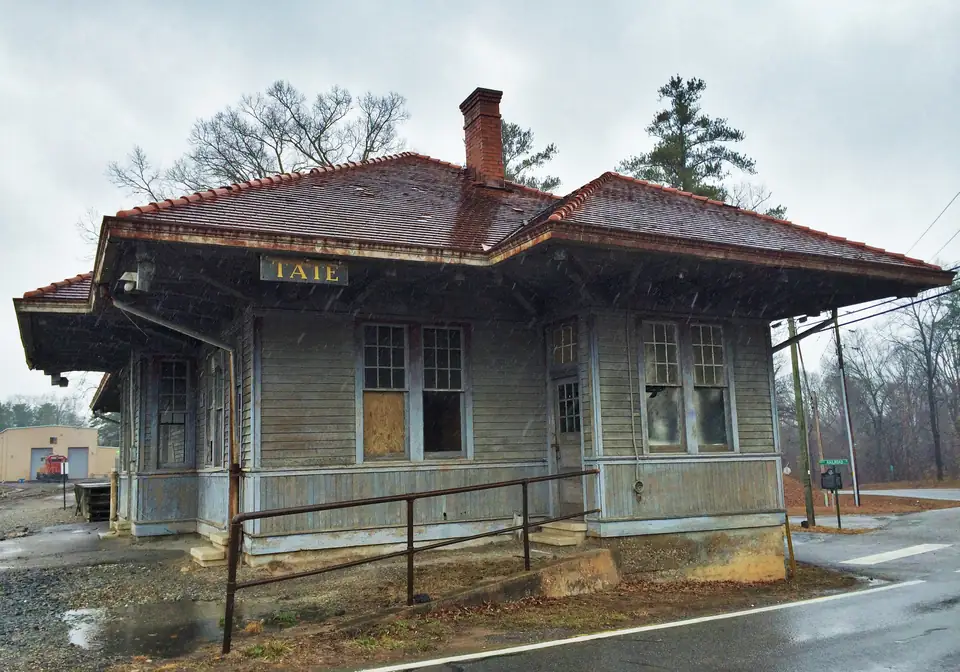A weathered clapboard railroad depot with a red tile roof and a TATE sign, seen in light snow.