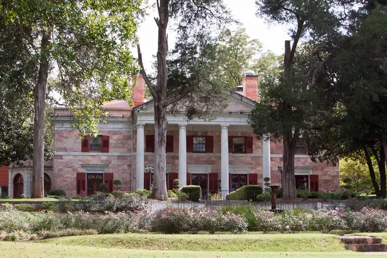 A two-story mansion faced in pink marble with a portico of four tall white columns, framed by shade trees.