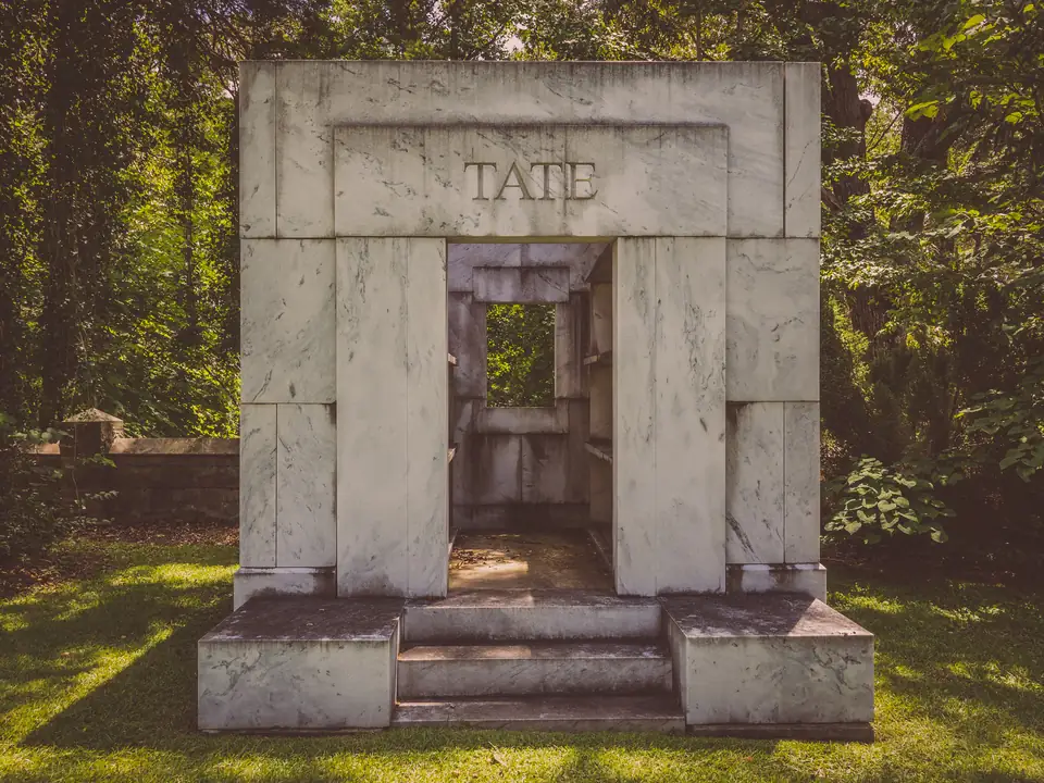 An open-fronted mausoleum of large veined white marble slabs with TATE carved in the lintel, in front of green woods.